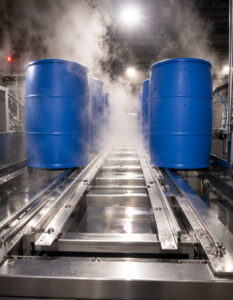 End view of an LS Industries Poly Washer with two plastic barrels positioned on the conveyor track, illustrating the automated material handling system for efficient barrel washing.