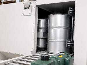 Exit side of a flow-through drum blaster showing a freshly blasted steel drum leaving the blasting chamber after surface preparation.