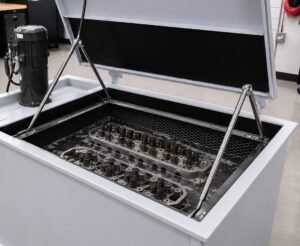 Interior view of a hot tank with a cylinder head partially submerged in heated water and cleaning chemicals, illustrating the immersion cleaning process for engine components.