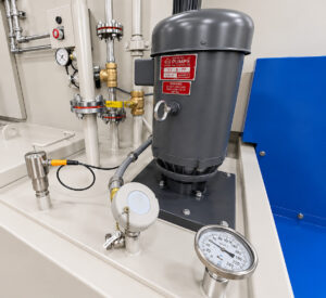 Industrial water pump on a monorail flow-through washer circulating cleaning solution throughout the wash system to power spray nozzles and maintain cleaning performance.