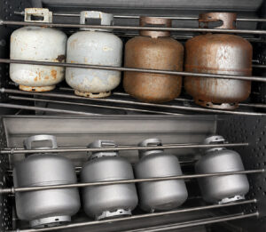 Side-by-side comparison of 20 lb propane tanks before and after processing through a propane batch tank blaster, showing restored clean surfaces after abrasive blasting.