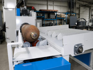 Cylinder positioned on conveyor wheels at the entrance of a flow-through cylinder blaster before entering the blasting chamber for processing.