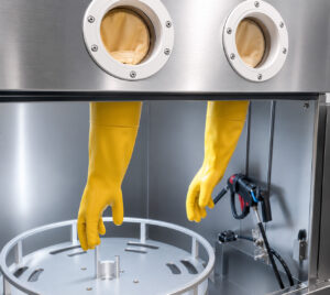 Protective rubber gloves mounted to the door of an industrial pressure washer, allowing operators to safely handle and clean parts inside the enclosed wash chamber.