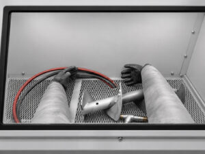 Operator's point-of-view through the viewing window of a glass bead blasting cabinet, showing a part inside the work chamber being prepared for blasting.