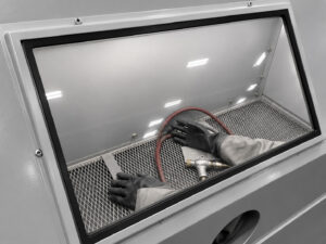 Protective blasting gloves visible through the viewing window of a glass bead blasting cabinet, ready for precision surface finishing operations.