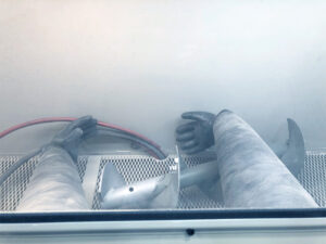 Point-of-view perspective of an operator working inside a glass bead blaster cabinet handling parts with built-in gloves during blasting.