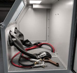 Interior view of a glass bead blasting cabinet through the access door, showing the work chamber, blast hose, gloves, and blasting area.