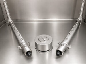 Underside view of a stainless steel washer table showing the spray nozzles and rotating table drive system responsible for delivering cleaning solution and rotating parts during the wash cycle.